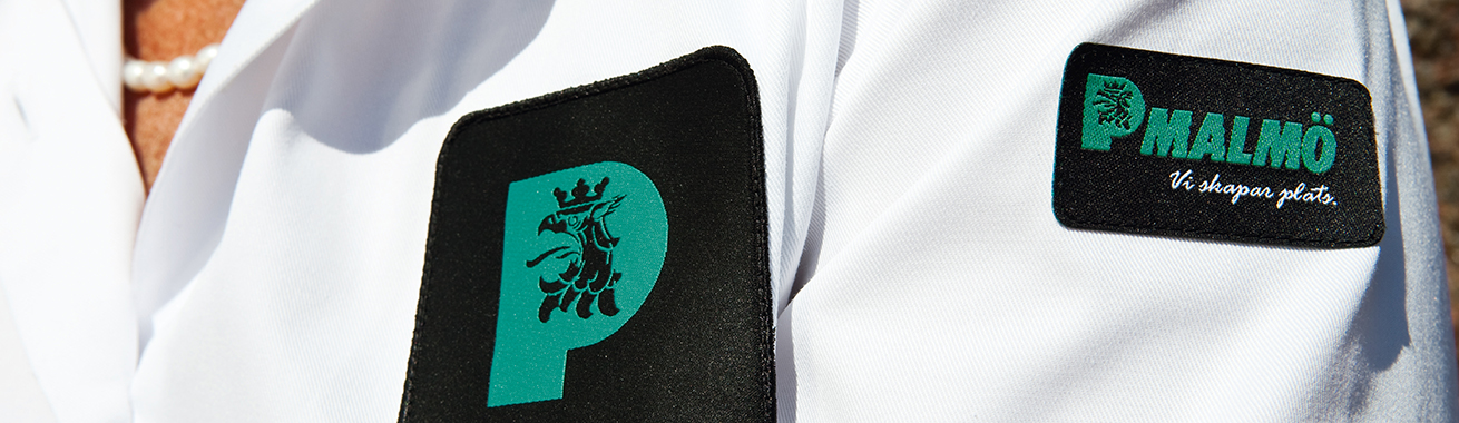 Närbild på en vit skjorta med vår logotype i grönt på svart botten på bröst och ärm 